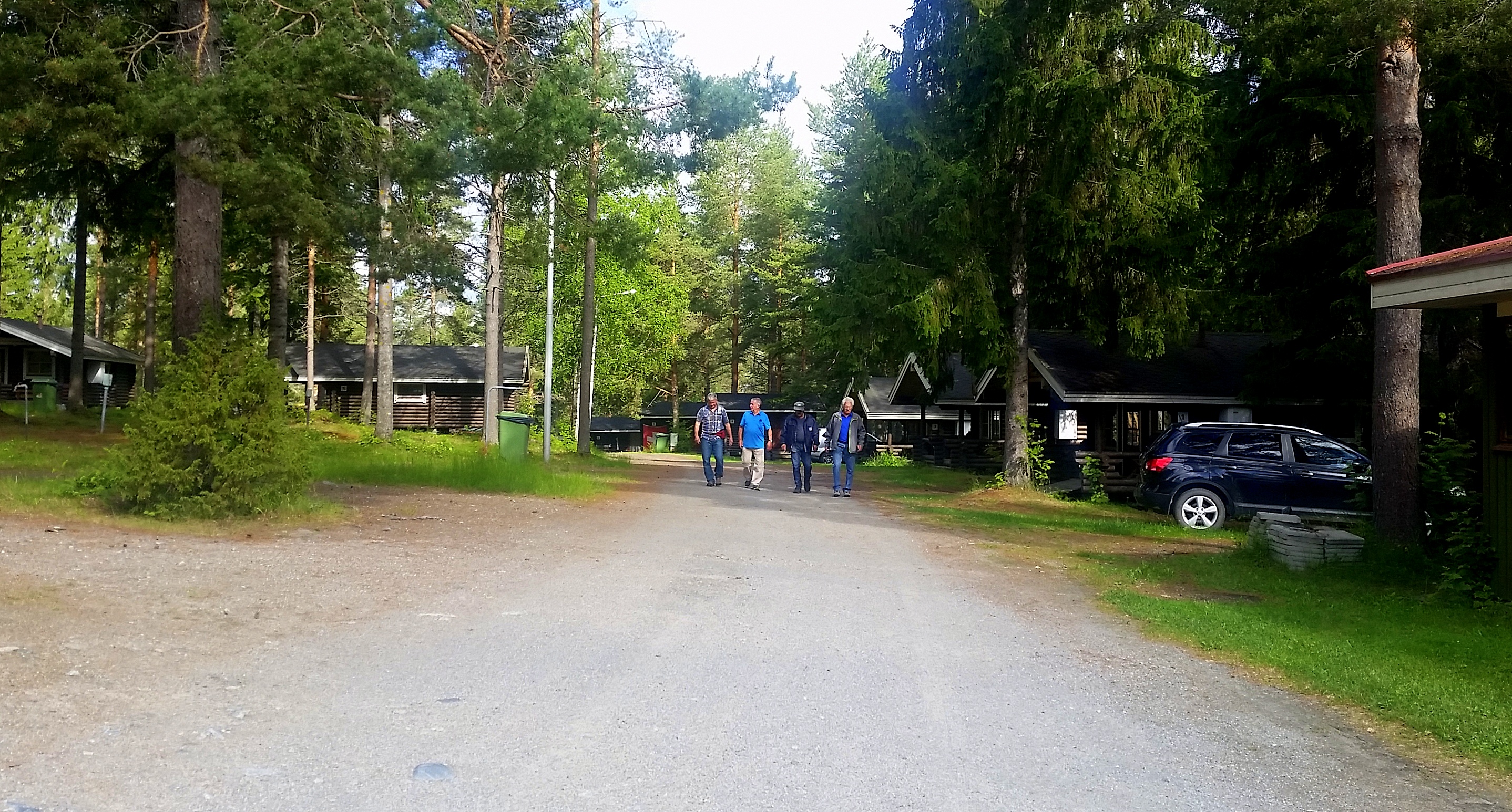 Einfahrt zum Campingplatz mit Kiesweg, Bäumen und Parkplatz mit Auto rechts, im Hintergrund Personen auf dem Weg.