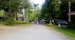 Einfahrt zum Campingplatz mit Kiesweg, Bäumen und Parkplatz mit Auto rechts, im Hintergrund Personen auf dem Weg.