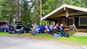 Musikgruppe mit Blasinstrumenten sitzt vor einem Campinghaus, neben geparkten Autos und unter Bäumen im grünen Campingbereich.