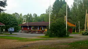 Restaurant-Terrasse auf einem Campingplatz mit Markise, Sitzplätzen und angrenzendem Weg, umgeben von Bäumen und Rasen.