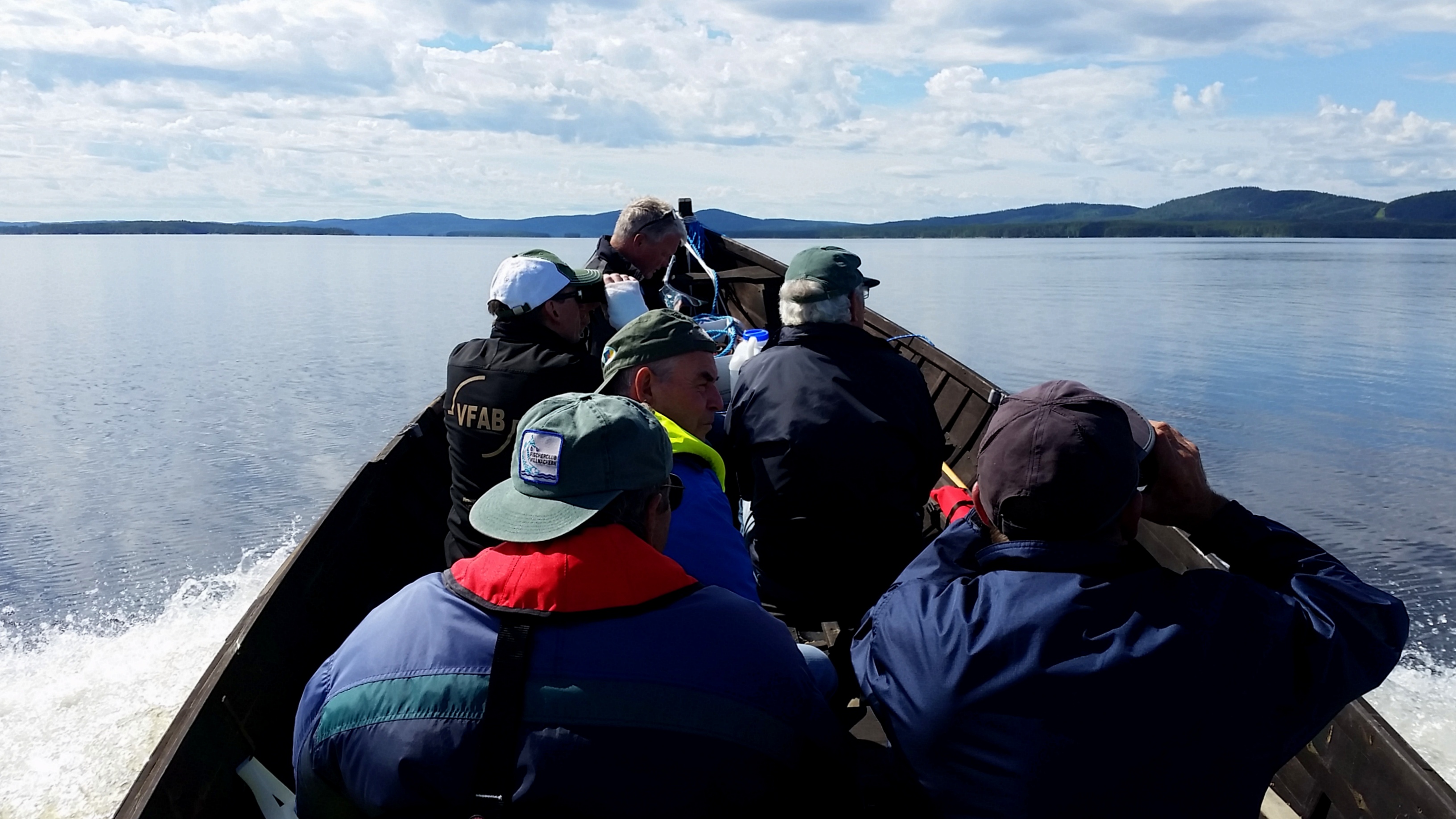 Boot mit mehreren Personen auf einem See, Wasser spritzt am Rand, im Hintergrund flache Ufer und Hügel.