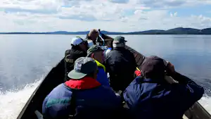 Boot mit mehreren Personen auf einem See, Wasser spritzt am Rand, im Hintergrund flache Ufer und Hügel.