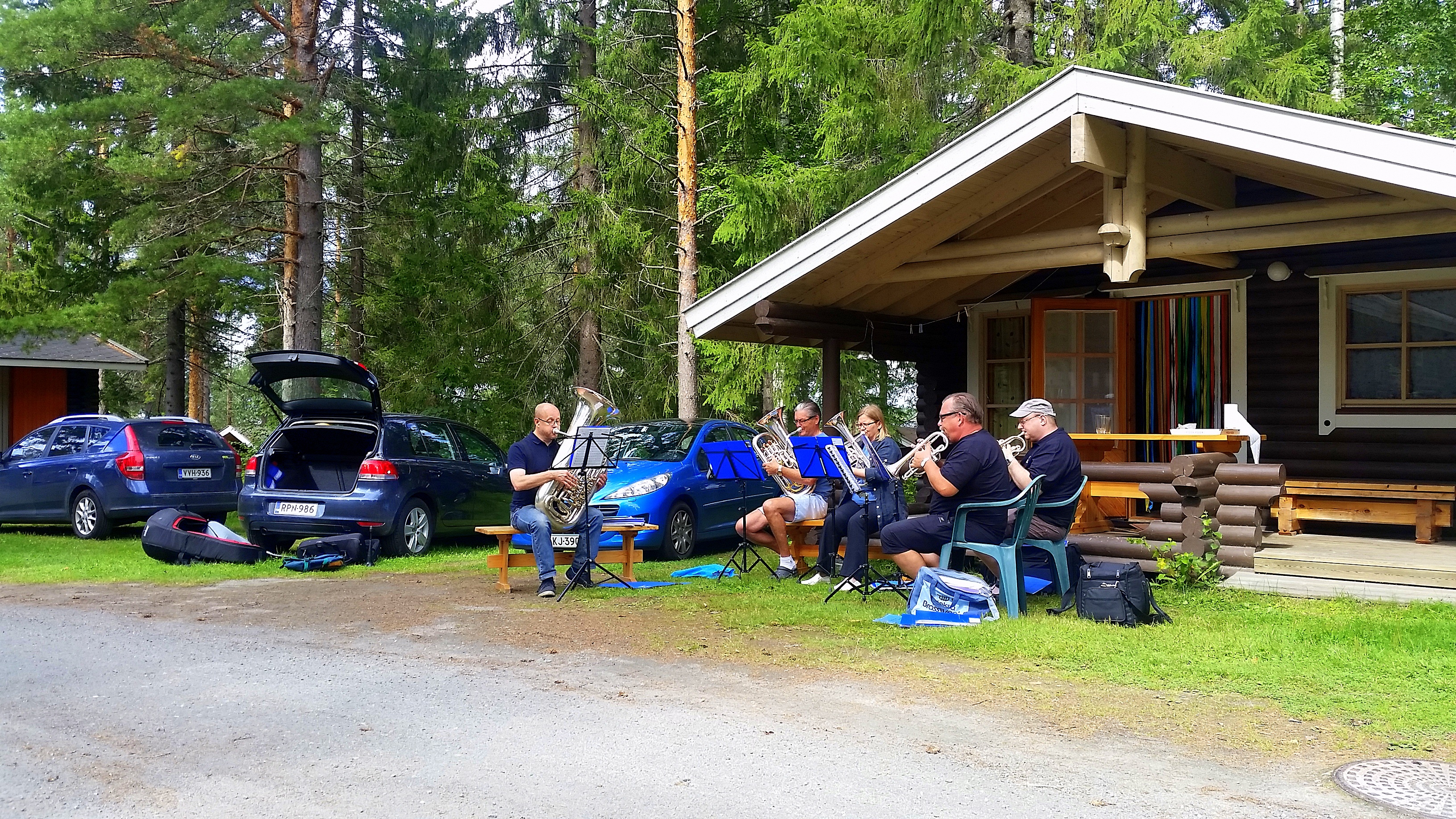 Campingplatz mit Wohnwagen und Terrasse am Holzhaus, daneben geparkte Autos und eine Gruppe am Tisch im Wald.