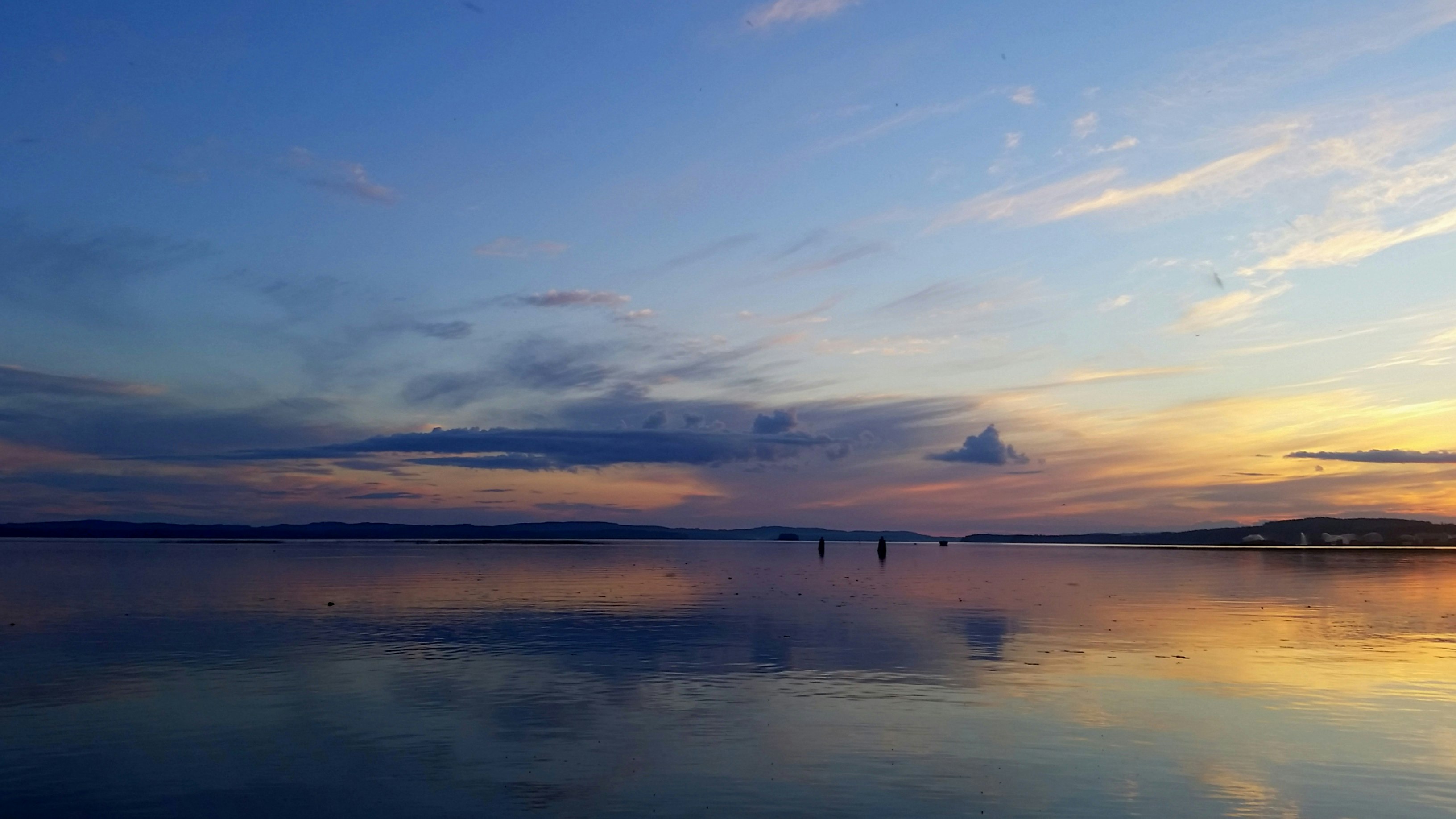 Ruhiger See bei Sonnenaufgang mit Spiegelung im Wasser, einzelne Boote und eine weite Uferlinie in der Ferne.