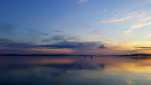 Ruhiger See bei Sonnenaufgang mit Spiegelung im Wasser, einzelne Boote und eine weite Uferlinie in der Ferne.