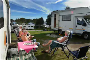 Wohnmobil auf dem Campingplatz, davor Tisch und Stühle auf dem Rasen, daneben weitere Fahrzeuge und Wohnwagen.