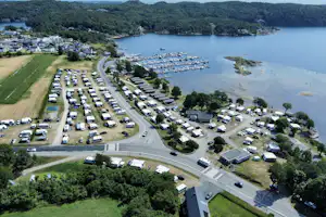 Luftaufnahme eines Campingplatzes am See mit Stellplätzen, Wohnmobilen, Wegen, Bäumen und Booten im Wasser.