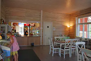 Camping-Shop mit Theke, Kühlschrank und weißen Tischen im Holzraum, daneben eine Tür und große Fenster.