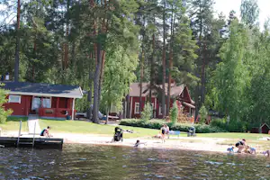 Rote Ferienhäuser am Seeufer im Wald, Rasenweg und Bäume im Hintergrund, Kinder spielen am Wasser.