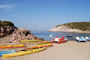 Strand mit mehreren Kajaks und Kanus auf dem Sand, im Hintergrund Boote und eine felsige Küste.