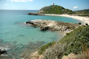 Blick auf eine Küstenbucht mit Sandstrand, klarem Meer und Felsen, im Hintergrund ein Hügel mit Turm.