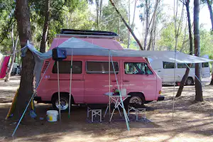 Rosa Wohnmobil mit Vorzelt und Campingstühlen am Stellplatz im Wald, daneben ein weiteres Fahrzeug.