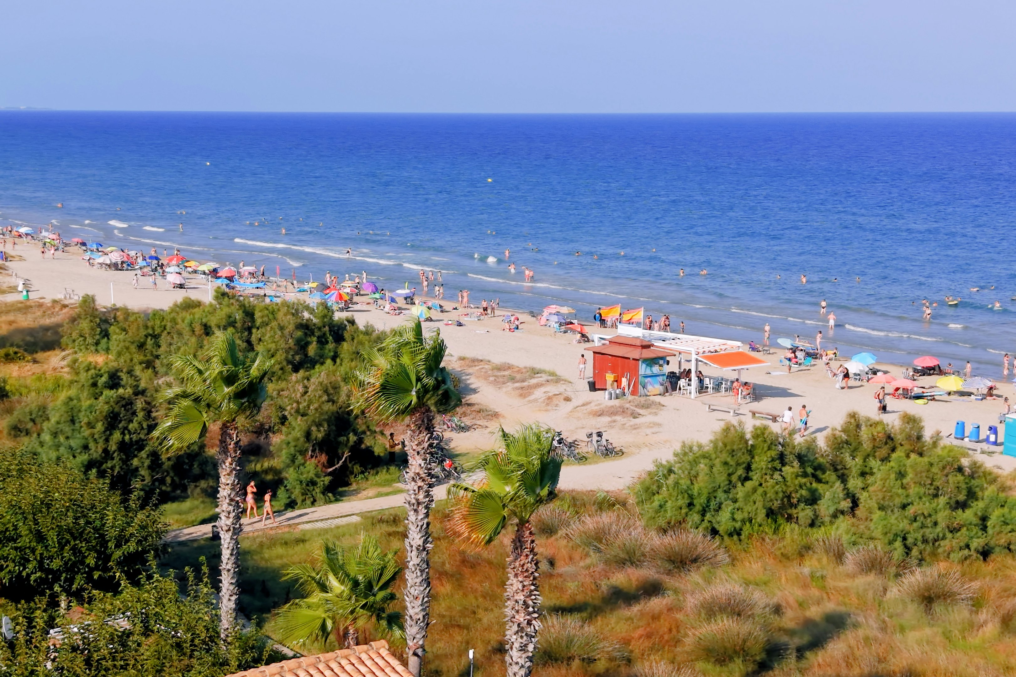 Blick auf einen Sandstrand am Meer mit Liegen, Sonnenschirmen und Promenade, umgeben von Bäumen und Hügeln.