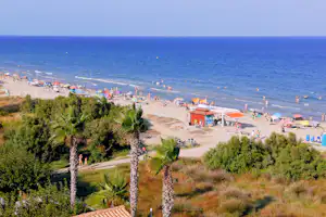 Blick auf einen Sandstrand am Meer mit Liegen, Sonnenschirmen und Promenade, umgeben von Bäumen und Hügeln.