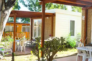 Terrasse am Mobilheim mit Holzüberdachung, weißen Gartenstühlen und Tisch, umgeben von Bäumen und Sträuchern.