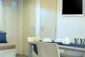 Wohnbereich mit Esstisch, Stühlen und Wasserkocher auf dem Tisch, daneben ein Sofa und Schranktüren.