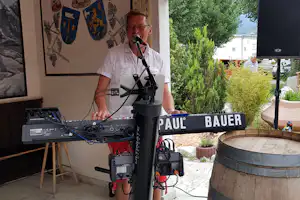 Mann spricht in ein Mikrofon vor einer Technik- und Lautsprecheranlage, im Hintergrund Hecken und Gebäude am Campingplatz.