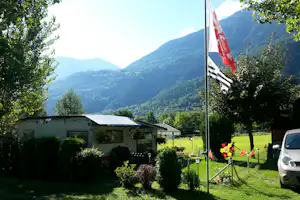 Campingplatz mit Wohnwagen, Fahnen am Mast und grüner Wiese; im Hintergrund Berge und bewaldete Hänge.