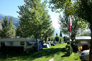 Wohnwagen und Zelte auf einem Campingplatz zwischen Bäumen, mit Autos und Bergen im Hintergrund.