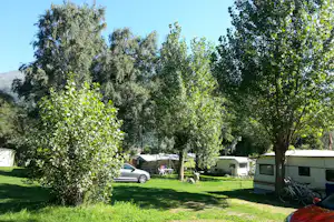 Campingplatz mit hohen Bäumen, Wohnwagen und Zelten im Hintergrund, dazu ein Auto und ein Fahrrad am Rand.