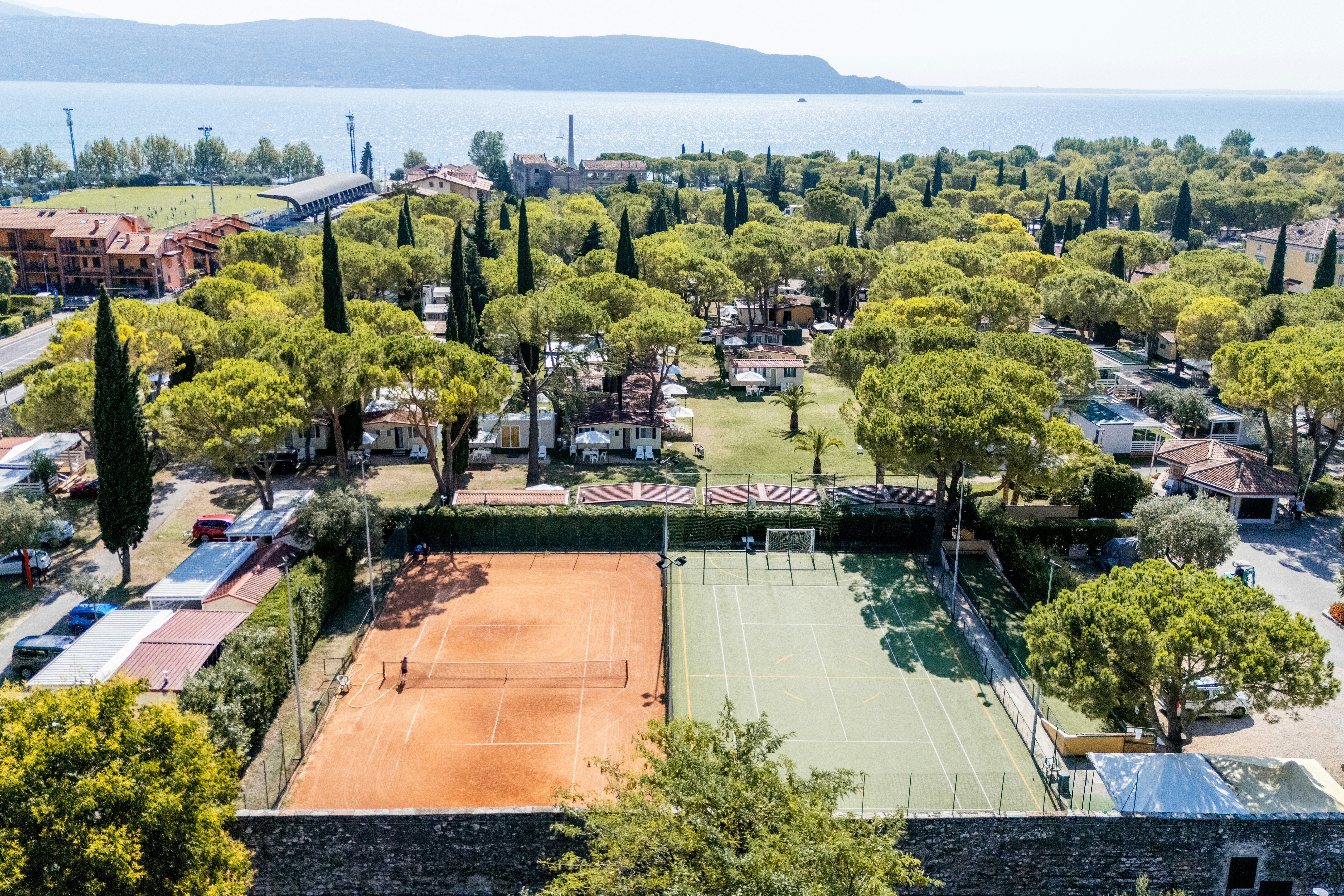 Campinganlage mit Tennisplatz, gepflegten Wegen und Bäumen, Wohnwagen und kleinen Gebäuden in einer grünen Umgebung.