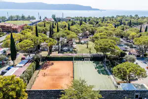 Campinganlage mit Tennisplatz, gepflegten Wegen und Bäumen, Wohnwagen und kleinen Gebäuden in einer grünen Umgebung.