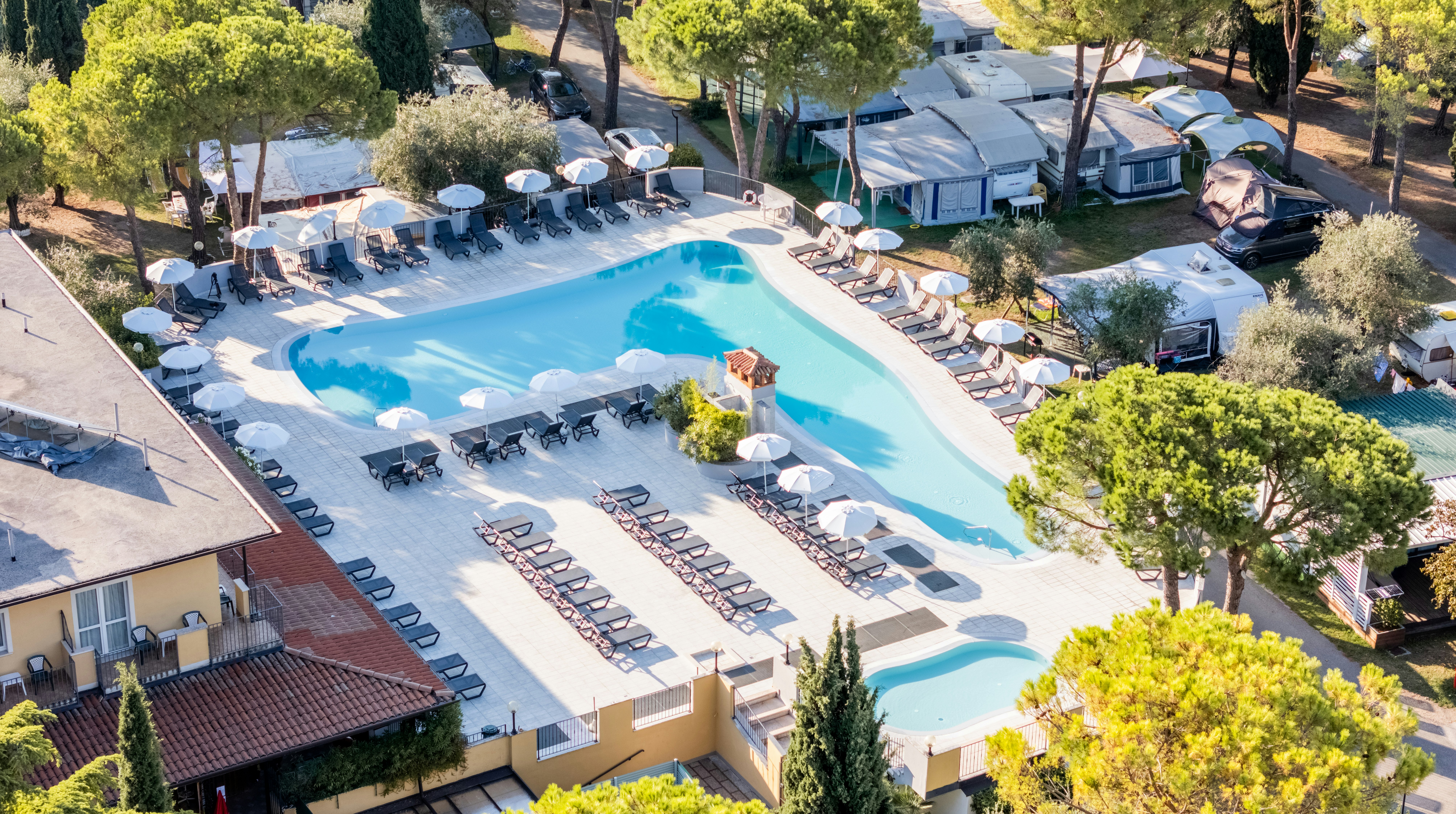 Großes Schwimmbecken mit Liegen am Campingplatz, daneben Zelte und Wohnwagen, umgeben von Bäumen und Wegen.