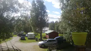Campingplatzweg mit Bäumen, geparktem Auto und Wohnwagen sowie einem kleinen roten Gebäude im Hintergrund.