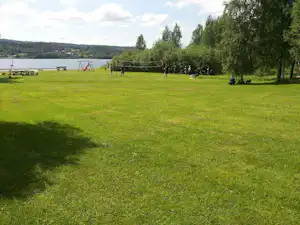 Grüne Campingwiese am See mit Bäumen, Spielgeräten und Sitzgelegenheiten am Ufer.