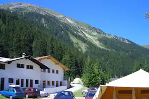 Campingplatz mit Zelt und geparkten Autos vor einem weißen Gebäude, umgeben von hohen Bergen und Nadelwald.