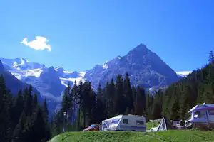 Campingplatz im Bergpanorama: Zelte und Wohnwagen auf einer Wiese, umgeben von Nadelbäumen und hohen Bergen.