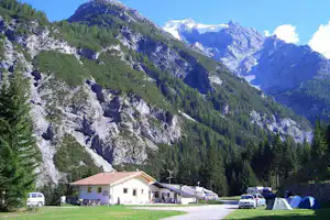 Campingplatz in den Alpen: Wohnhaus, Zelte und geparkte Autos am Weg, umgeben von Tannen und hohen Bergen.