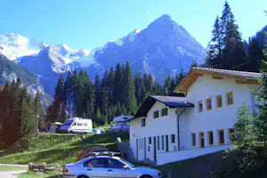 Campinghaus mit Parkplatz und Autos, im Hintergrund hohe Berge und Nadelwald in den Alpen.