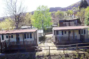 Campingbungalows mit Holzterrassen und Sichtschutz, gepflasterter Weg dazwischen, Bäume und Hügel im Hintergrund.