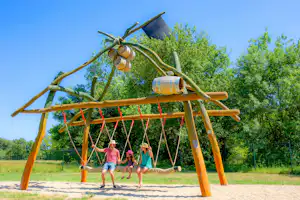 Spielplatz auf dem Campingplatz: Holzgestell mit Schaukeln, Kinder laufen im Hintergrund vor grünen Bäumen.