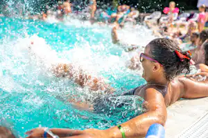 Kind und Familie planschen im Campingpool, Wasser spritzt, am Beckenrand sitzen weitere Personen.