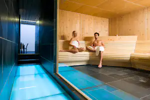 Sauna mit Holzbank und blau beleuchtetem Tauchbecken, zwei Personen sitzen und liegen auf den Liegen.