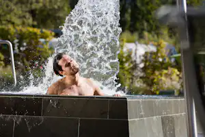 Mann unter einem Wasserstrahl aus der Dusche, Spritzwasser und Hintergrund mit Bäumen und Campinganlage.