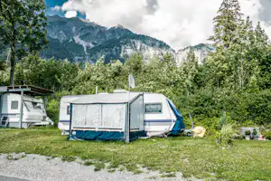 Wohnwagen und Vorzelt auf dem Campingplatz, umgeben von Bäumen, mit Blick auf hohe Berge und Wolken.
