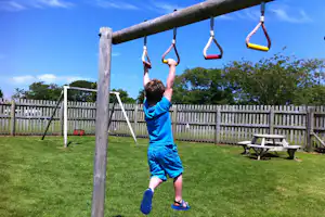 Kind schwingt an einer Schaukel auf dem Spielplatz am Campingplatz, mit Rasen, Holzzaun und Liege auf der Wiese.