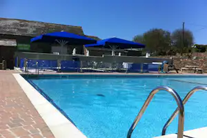 Blaues Camping-Poolbecken mit Edelstahlleiter, Ziegelweg und Liegen unter Sonnenschirmen neben dem Gebäude.