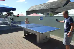 Tischtennisplatte auf dem Campingplatz, daneben Stühle und Sonnenschirm; im Hintergrund Gebäude und Zeltplanen.