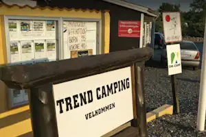 Schild „TREND CAMPING“ mit „VELKOMMEN“ am Eingang, daneben Infotafel und geparkter Pkw vor dem Gebäude.