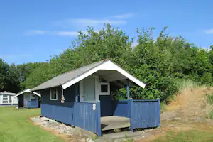Blaues Holzhaus mit Veranda und Eingang, umgeben von Bäumen und Wiese auf einem Campingplatz.