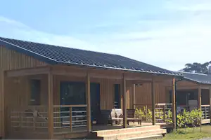 Holzgebäude mit Satteldach und Solarpanelen, Terrasse mit Stühlen und Geländer, im Hintergrund Bäume und Himmel.