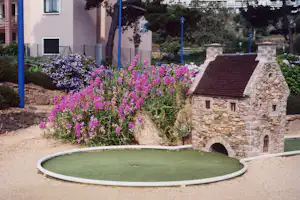 Kleiner Steinbrunnen mit rundem Becken, umgeben von bunten Blumenbeeten und Hecken vor einem Campinggebäude.