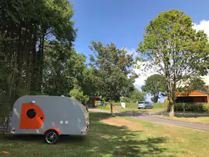 Kleiner Wohnwagen auf einem Campingplatz am Grasweg, umgeben von Bäumen und einem Gebäude im Hintergrund.