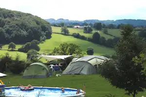 Zelte auf einem Campingplatz auf einer grünen Wiese, im Vordergrund ein Planschbecken und Blick auf Hügel und Bäume.