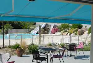 Terrasse mit Stühlen und Sonnenschirm am Campingpool, im Hintergrund Wasserbecken, Rutsche und gepflegte Blumenbeete.