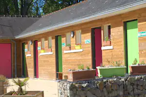 Holzgebäude mit bunten Türen und WC-Schildern, davor Pflanzkübel und ein Weg am Campingplatz.